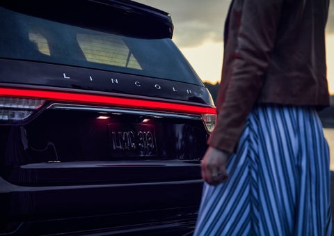 A person is shown near the rear of a 2024 Lincoln Aviator® SUV as the Lincoln Embrace illuminates the rear lights | Vista Lincoln Woodland Hills in Woodland Hills CA