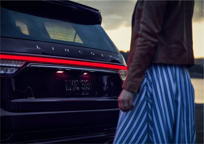 A person is shown near the rear of a 2023 Lincoln Aviator® SUV as the Lincoln Embrace illuminates the rear lights | Vista Lincoln Woodland Hills in Woodland Hills CA
